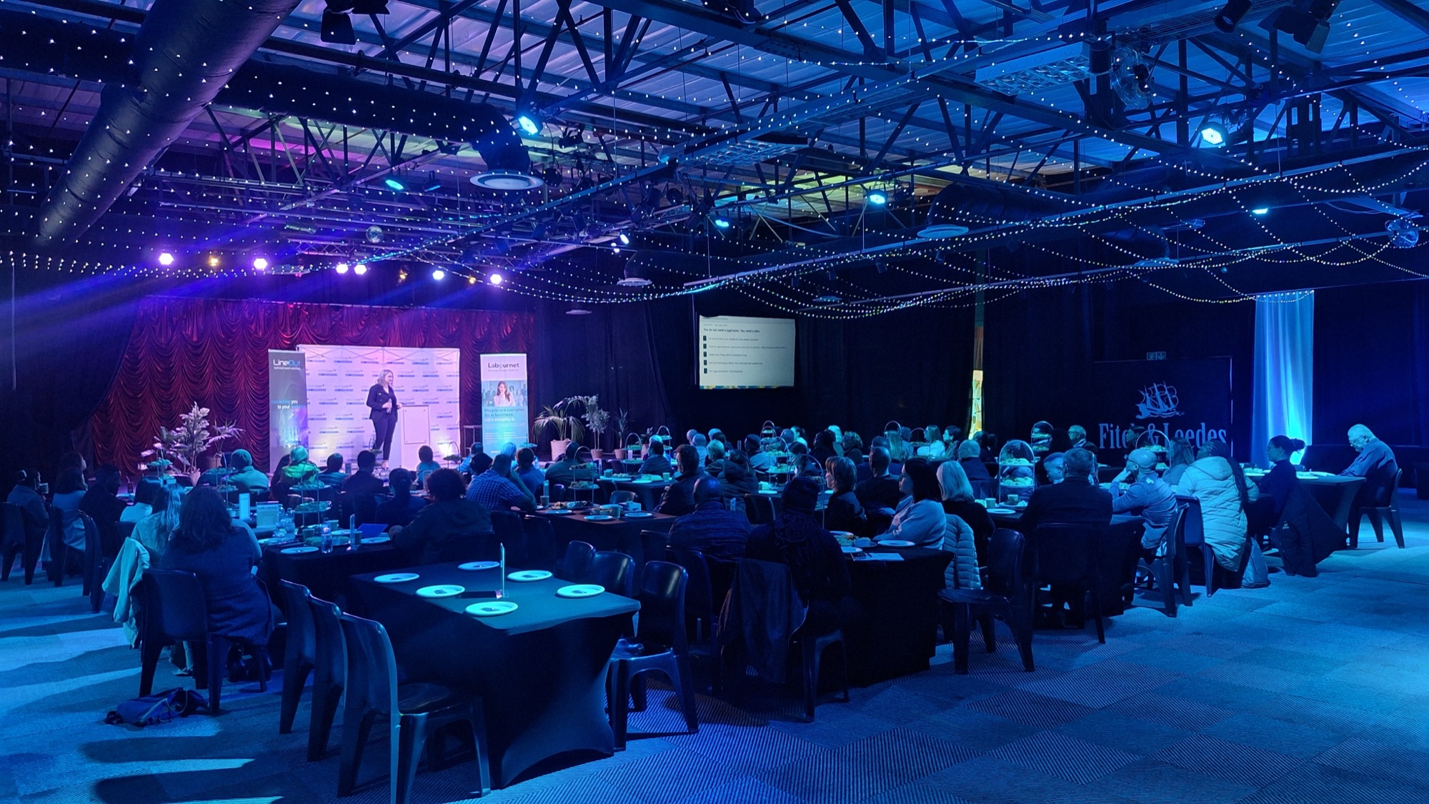An audience seated at tables watching a speaker at Centrestage@Baywest